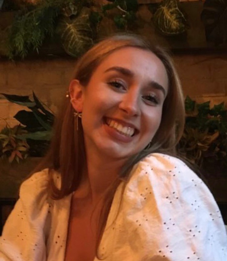 Portret uśmiechniętej młodej kobiety z blond włosami, w białej bluzce z ażurowymi detalami, na tle roślin i ceglanej ściany.