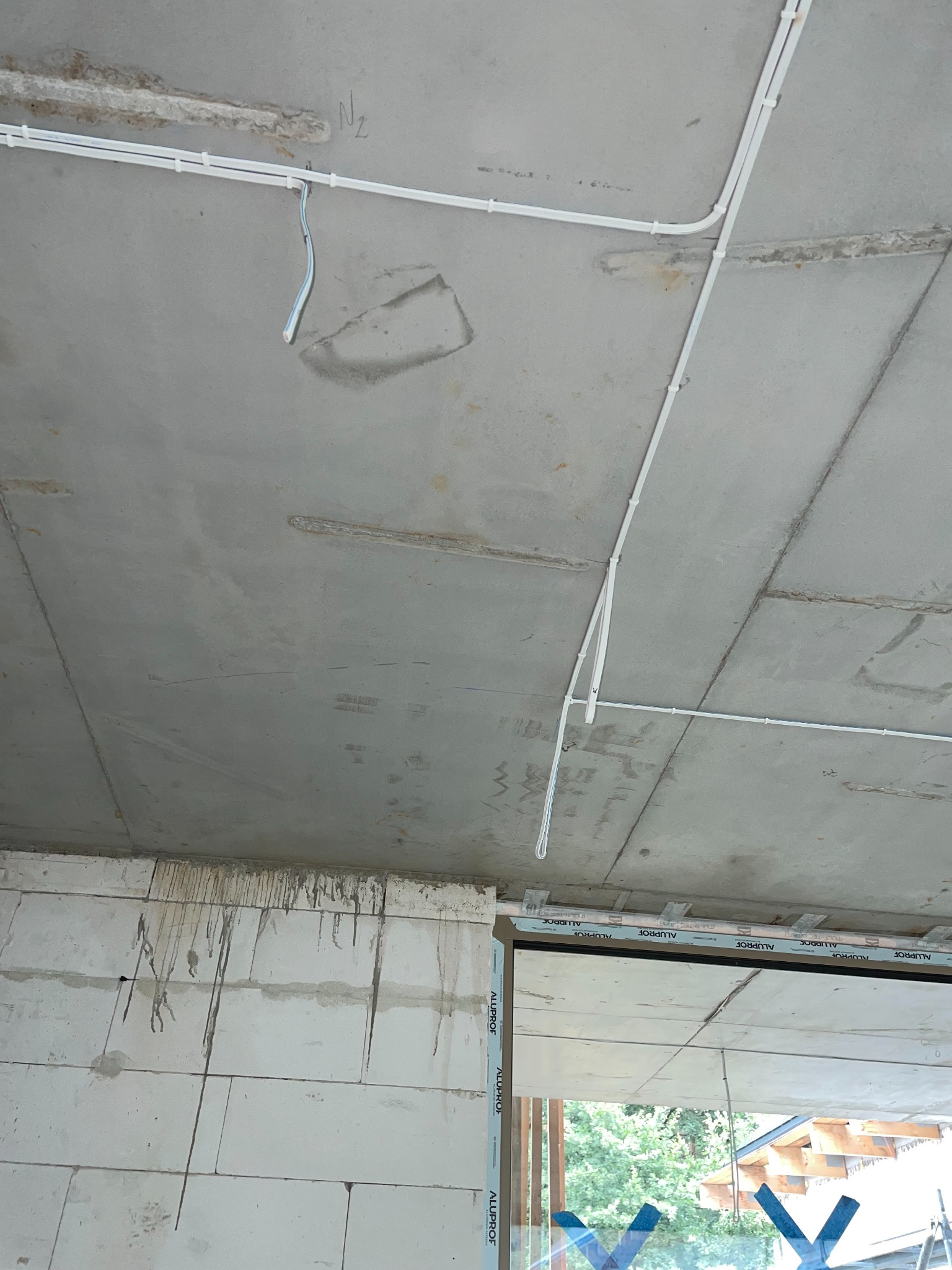 Białe kable elektryczne prowadzone po surowym, betonowym suficie w nowym budynku, widoczne fragmenty ścian z białego betonu komórkowego i okna z profili aluminiowych.