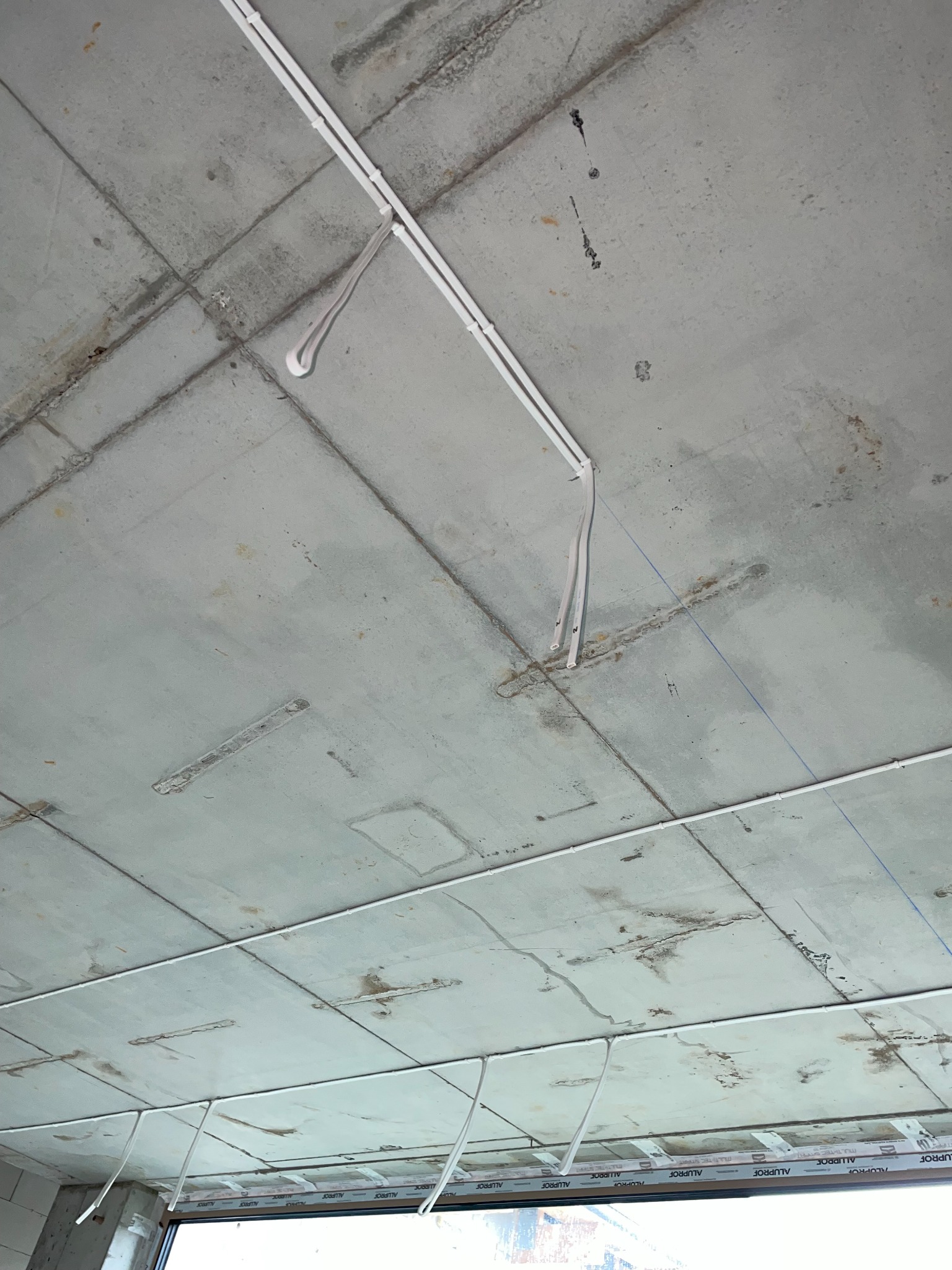 Surowy betonowy sufit z zamontowanymi białymi rurkami instalacji elektrycznej, widoczne kable zwisające w dół, okno w tle.