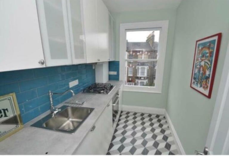 Wąska kuchnia z niebieską glazurą ułożoną w cegiełkę, białe szafki wiszące, szara zabudowa, wzorzysta podłoga i okno z widokiem na budynki.