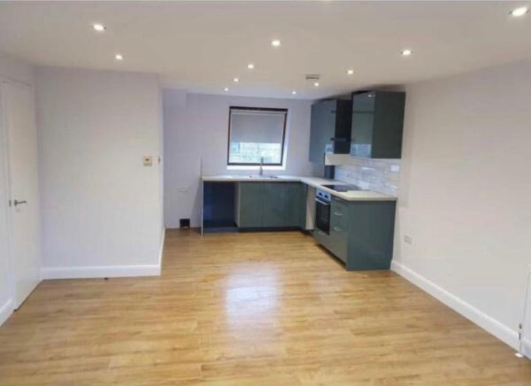 Jasne wnętrze po remoncie: kuchnia z zabudową w odcieniach szarości, drewniana podłoga, białe ściany i sufit z punktowym oświetleniem. Widoczne okno z roletą i fragment drzwi.