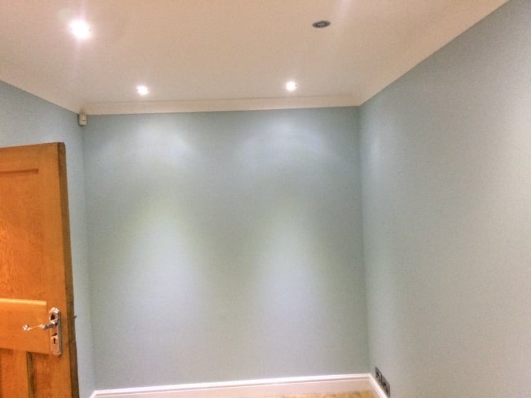 Świeżo pomalowane wnętrze pokoju w odcieniach błękitu, z drewnianymi drzwiami po lewej stronie i listwami przypodłogowymi. Widoczne punkty oświetleniowe w suficie.