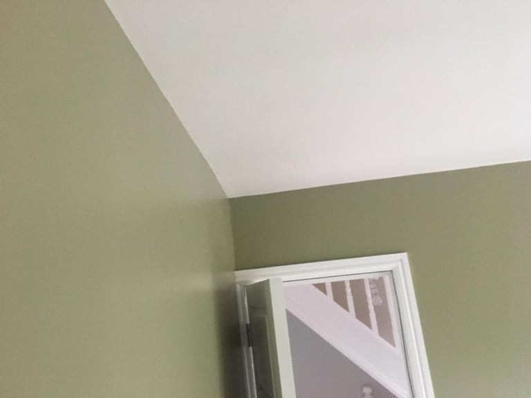 Świeżo pomalowane ściany w odcieniach oliwkowej zieleni i biały sufit, widoczny fragment białych schodów za otwartymi drzwiami.