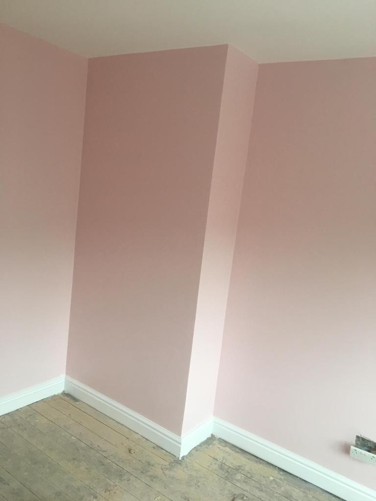 Różowe ściany po świeżym pomalowaniu z białymi listwami przypodłogowymi i surową drewnianą podłogą.