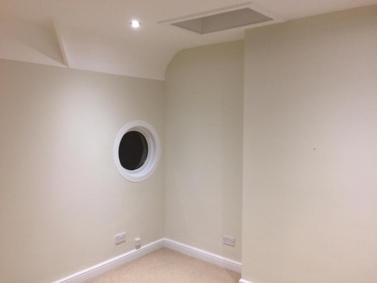 Jasne wnętrze po malowaniu: ściany w odcieniach beżu, okrągłe okno z białym obramowaniem, widoczne gniazdka elektryczne przy podłodze.