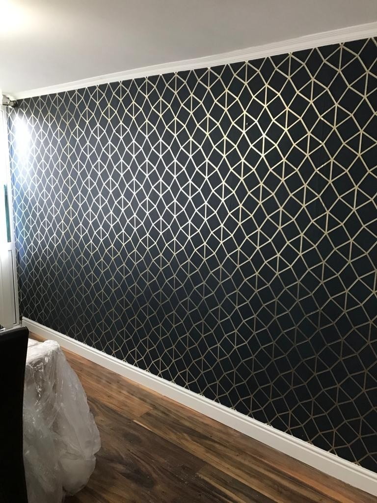 Ściana w pokoju pokryta ciemną tapetą ze złotym geometrycznym wzorem, widoczna listwa przypodłogowa i drewniana podłoga.