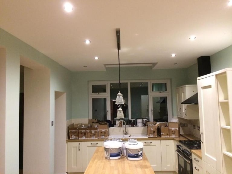 Kuchnia w trakcie odświeżania: ściany pomalowane na delikatny odcień zieleni, widoczne puszki z farbą na drewnianym blacie wyspy kuchennej, kartony z akcesoriami malarskimi na blacie przy zlewie...