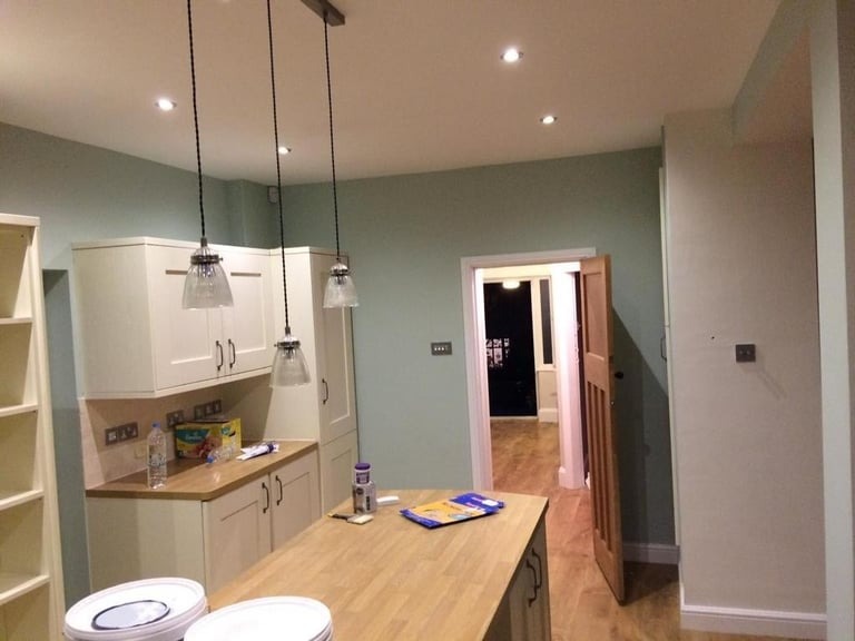 Świeżo pomalowane na pastelowo zielono ściany w kuchni w Gdańsku, z drewnianym blatem i wiszącymi lampami, widoczne puszki z farbą na wyspie kuchennej.