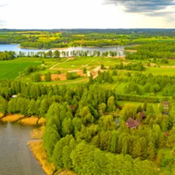 Panoramiczny widok z lotu ptaka na jezioro otoczone lasami i polami uprawnymi, z widocznymi domami z czerwonymi dachami, w słoneczny dzień z chmurami.