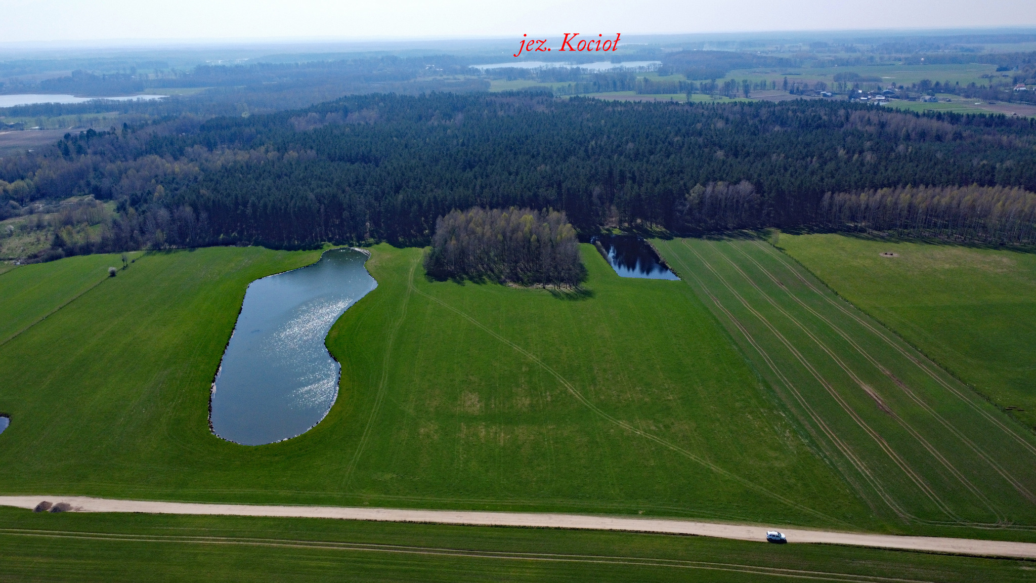 Panoramiczny widok z lotu ptaka na zielone pola uprawne z dwoma małymi jeziorami i lasem w tle, z widoczną polną drogą i małym samochodem