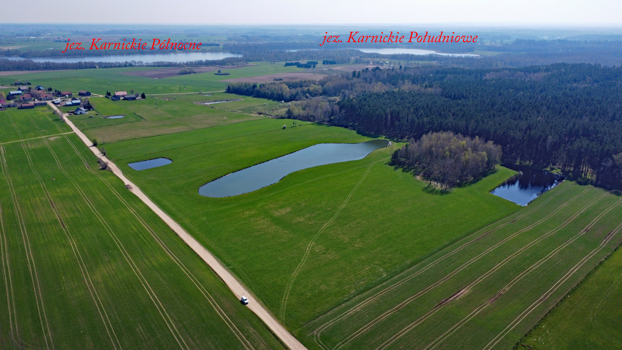 Panoramiczny widok z lotu ptaka na zielone pola uprawne, jeziora Karnickie Północne i Południowe oraz las, z białą furgonetką jadącą polną drogą.