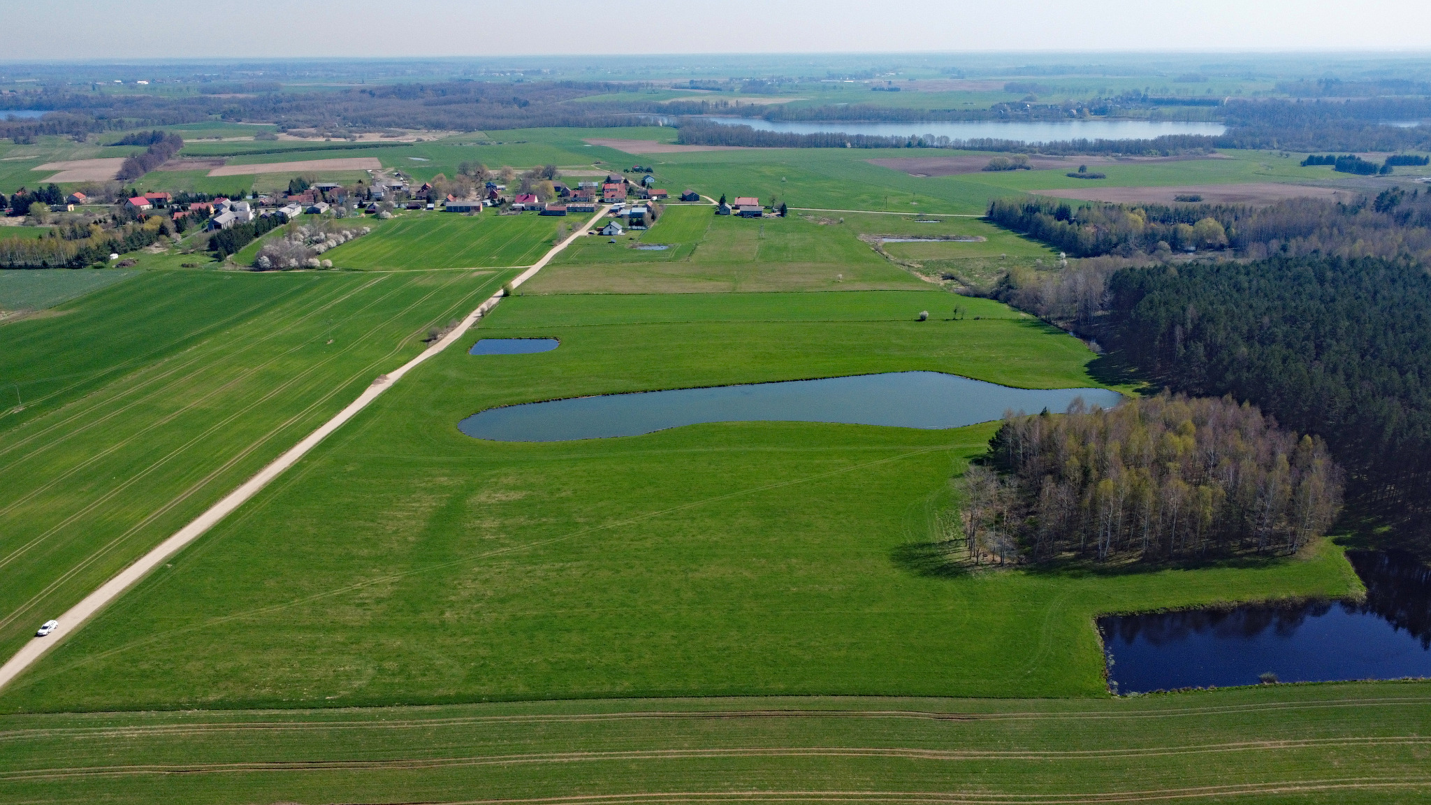 Rozległy krajobraz z lotu ptaka ukazujący zielone pola, małe jeziora, lasy i wieś w oddali, z białym samochodem jadącym polną drogą.