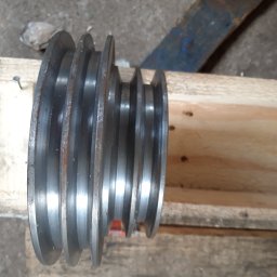 Ślusarski Zakład Produkcyjno-Handlowy Tadeusz Jarończyk - Metalowa, wielorowkowa rolka pasowa leżąca na drewnianych deskach, widoczne ślady korozji i zabrudzenia.