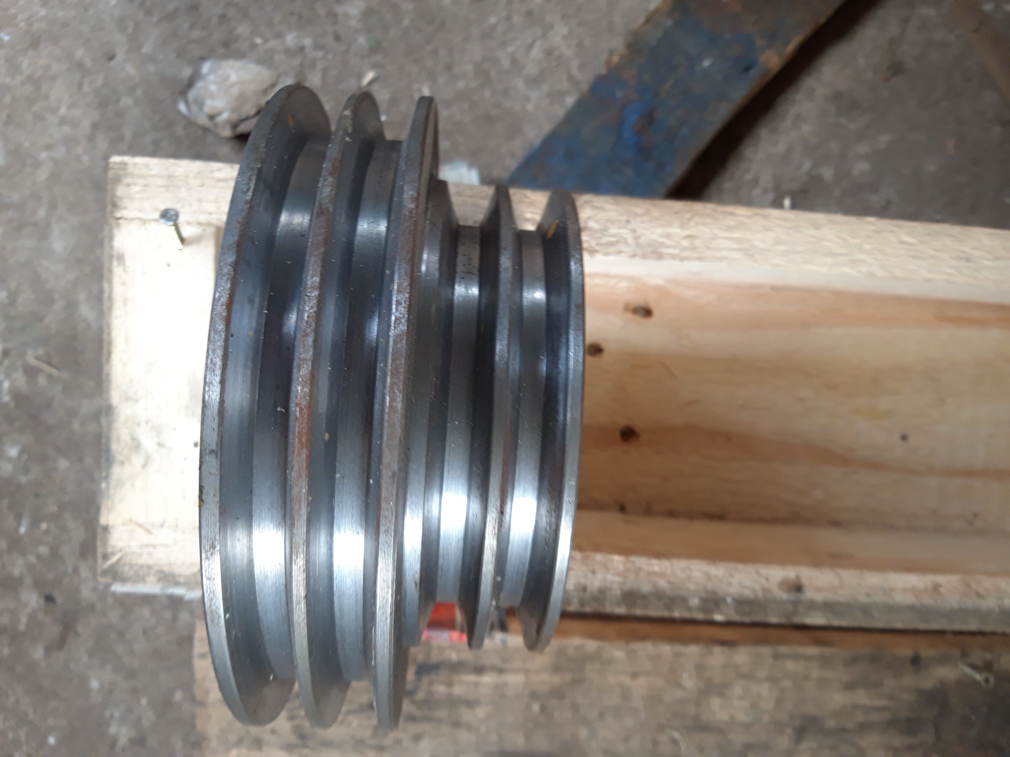 Metalowa, wielorowkowa rolka pasowa leżąca na drewnianych deskach, widoczne ślady korozji i zabrudzenia.