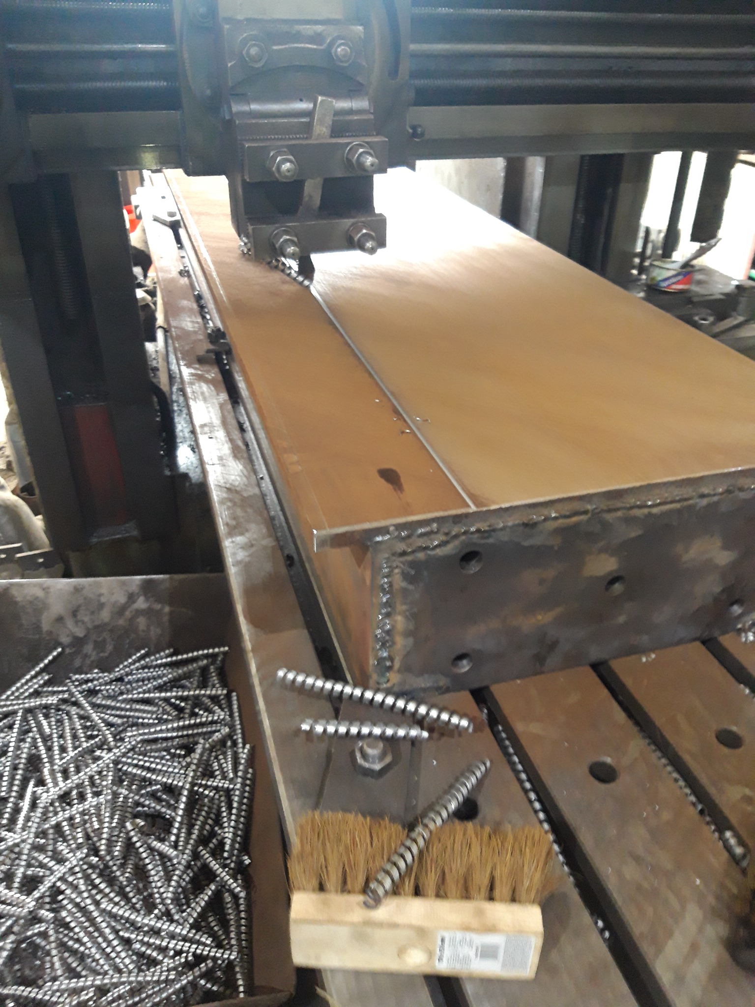 Frezarka pionowa obrabia metalową płytę, w tle widoczny pojemnik z metalowymi wiórami oraz szczotka do czyszczenia.