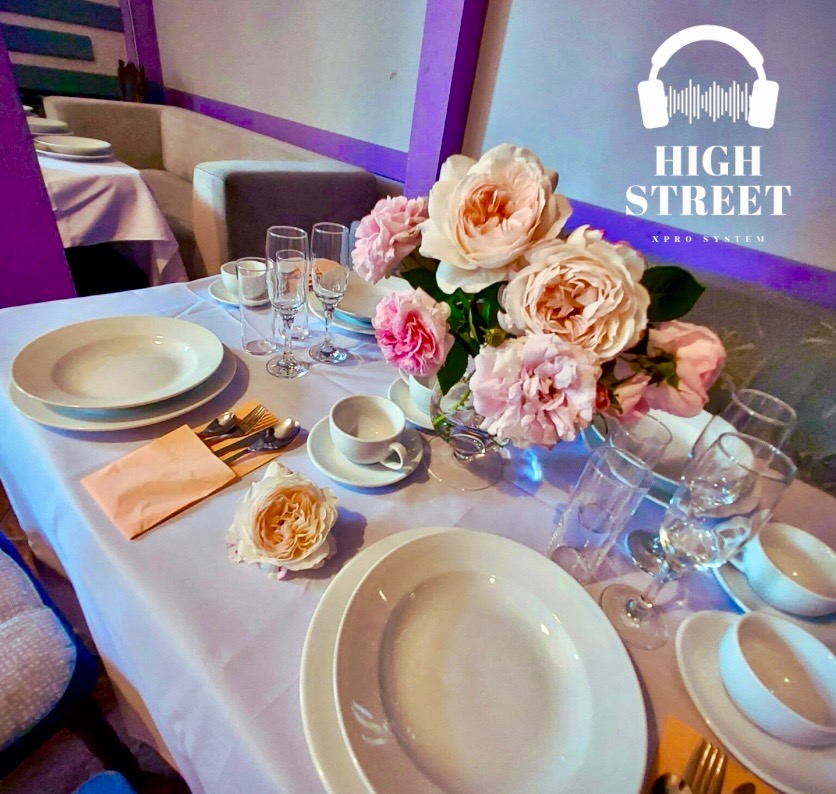 Elegancko nakryty stół z białym obrusem, porcelanową zastawą, srebrnymi sztućcami i bukietem różowych róż w szklanym wazonie, przygotowany na przyjęcie lub uroczystość, z widocznym logo firmy 'High...