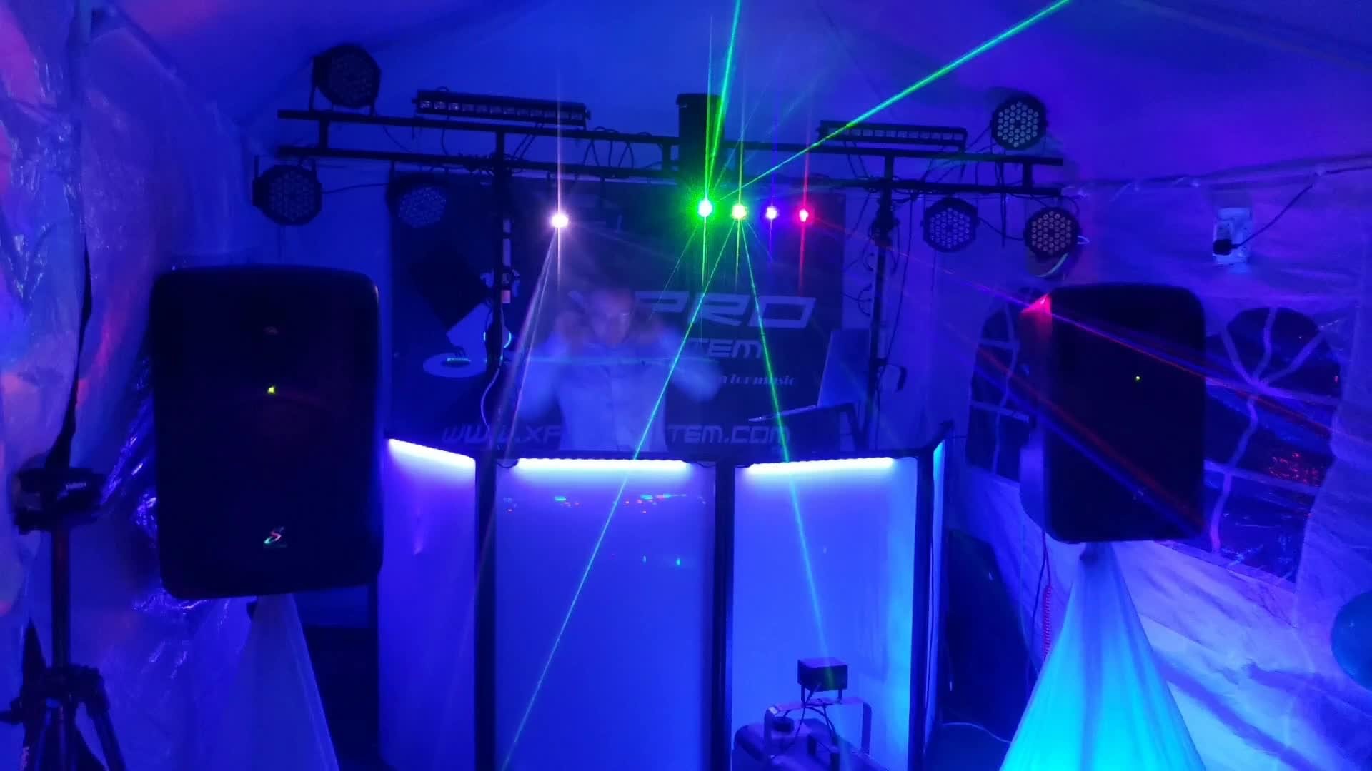 DJ za konsoletą oświetloną niebieskim światłem, z laserami przecinającymi przestrzeń, głośniki po bokach, tło z folii imitujące okno.