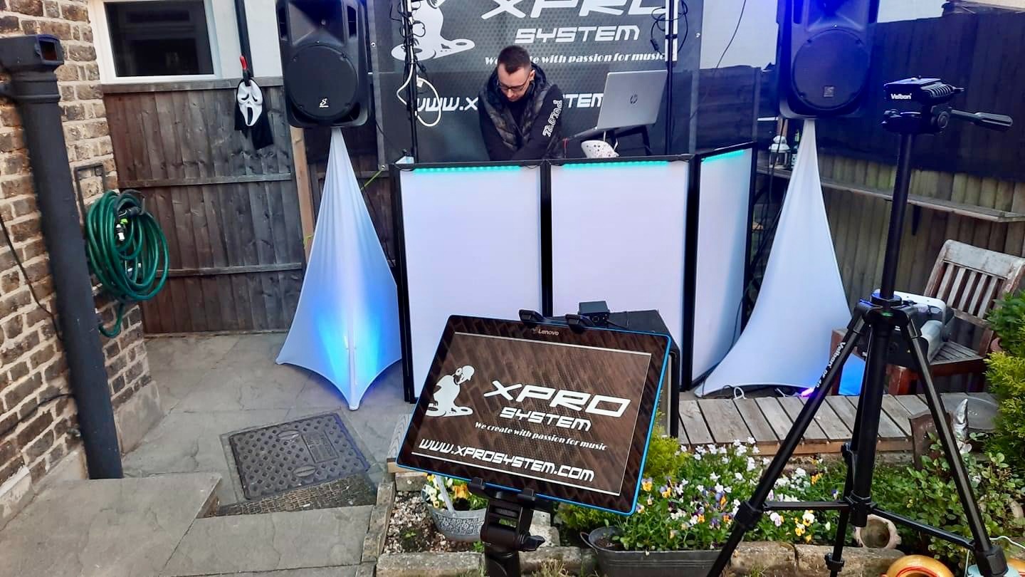 DJ za konsolą z laptopem i sprzętem nagłośnieniowym, widoczne kolumny, oświetlenie LED, maska Scream zawieszona na kolumnie, na pierwszym planie tablica z logo 'XPRO SYSTEM', tło stanowi ogród.