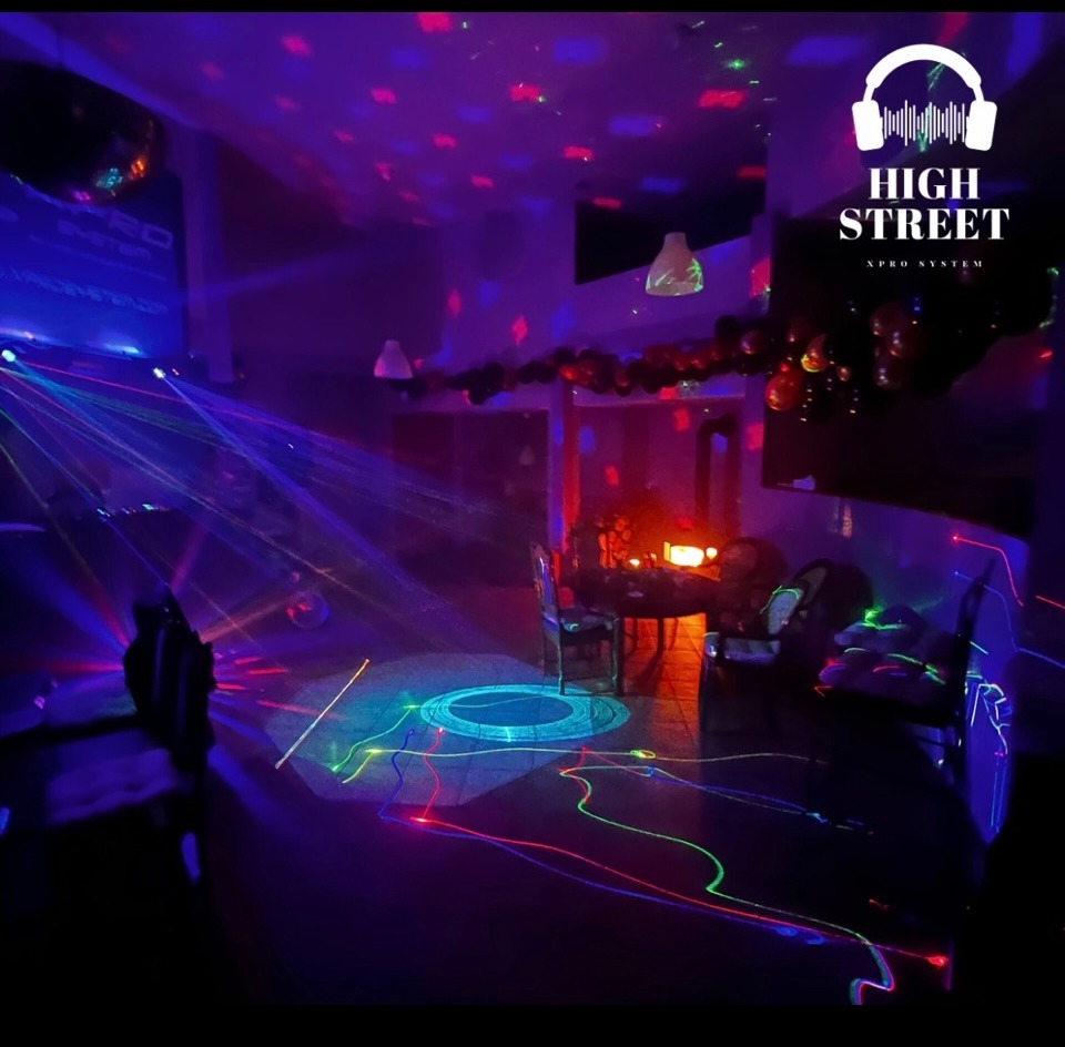 Sala klubowa oświetlona kolorowymi laserami, z dekoracjami balonowymi i stołem z krzesłami w tle, logo firmy nagłośnieniowej w rogu.