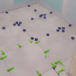 Układanie jasnych płytek podłogowych w narożnej łazience z użyciem poziomujących klipsów i klinów; widoczne narzędzia glazurnicze.