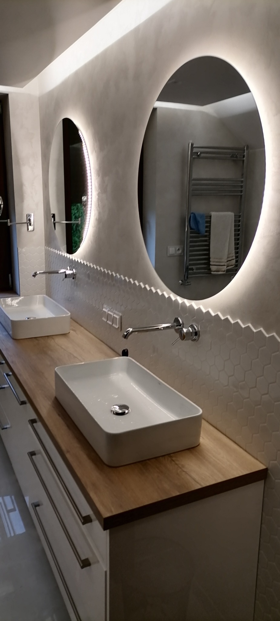 Nowoczesna łazienka z dwoma prostokątnymi umywalkami nablatowymi, drewnianym blatem, białymi szafkami z szufladami oraz okrągłymi lustrami podświetlanymi LED, wykończona drobną, białą mozaiką...