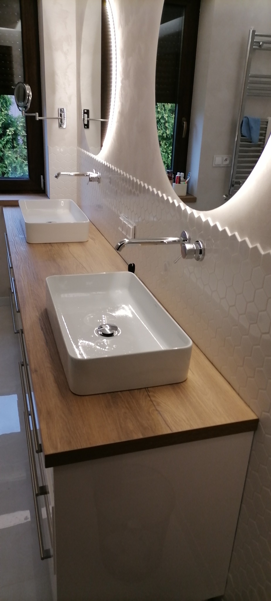 Nowoczesna łazienka w Rzeszowie z dwiema prostokątnymi umywalkami nablatowymi na drewnianym blacie, podświetlanym lustrem o nieregularnym kształcie i heksagonalnymi płytkami na ścianie.