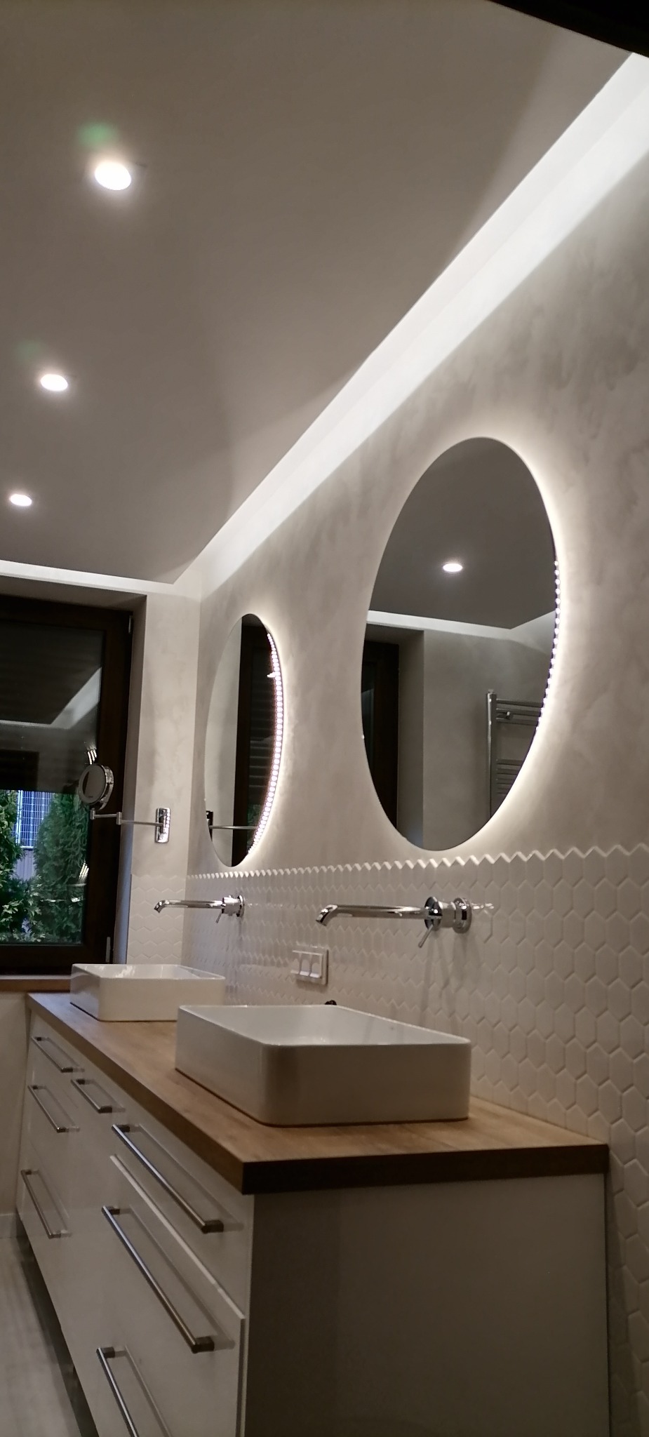 Nowoczesna łazienka z dwoma umywalkami nablatowymi, drewnianym blatem, białymi szafkami z podłużnymi uchwytami oraz dwoma okrągłymi lustrami z podświetleniem LED, heksagonalne płytki na ścianie.