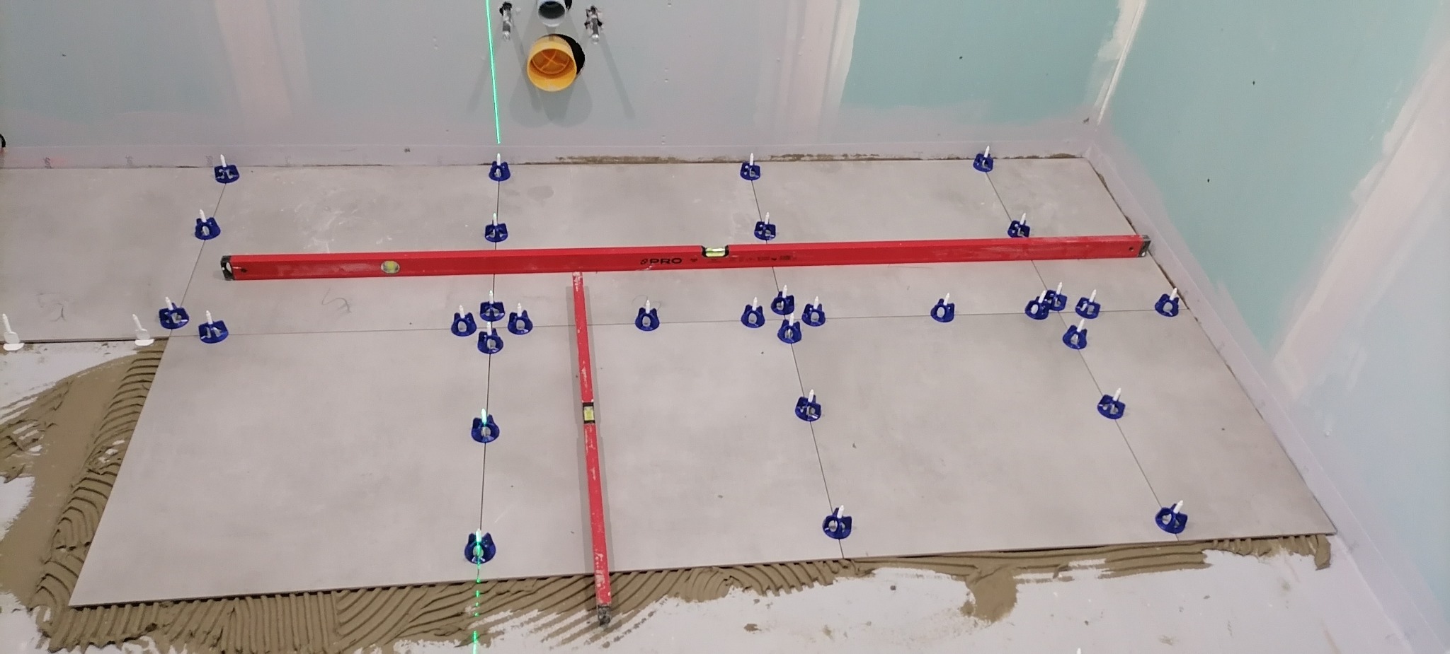 Układanie jasnoszarych płytek podłogowych w łazience, widoczne poziomice i system poziomowania płytek z niebieskimi klipsami, ściany przygotowane pod malowanie.