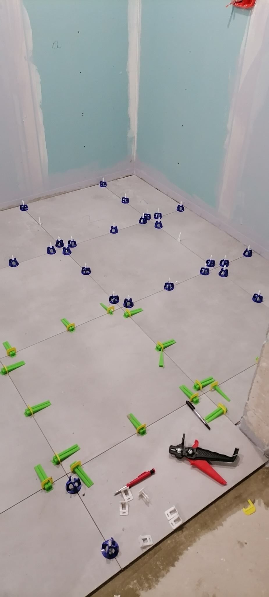 Układanie jasnych płytek podłogowych w narożnej łazience z użyciem poziomujących klipsów i klinów; widoczne narzędzia glazurnicze.