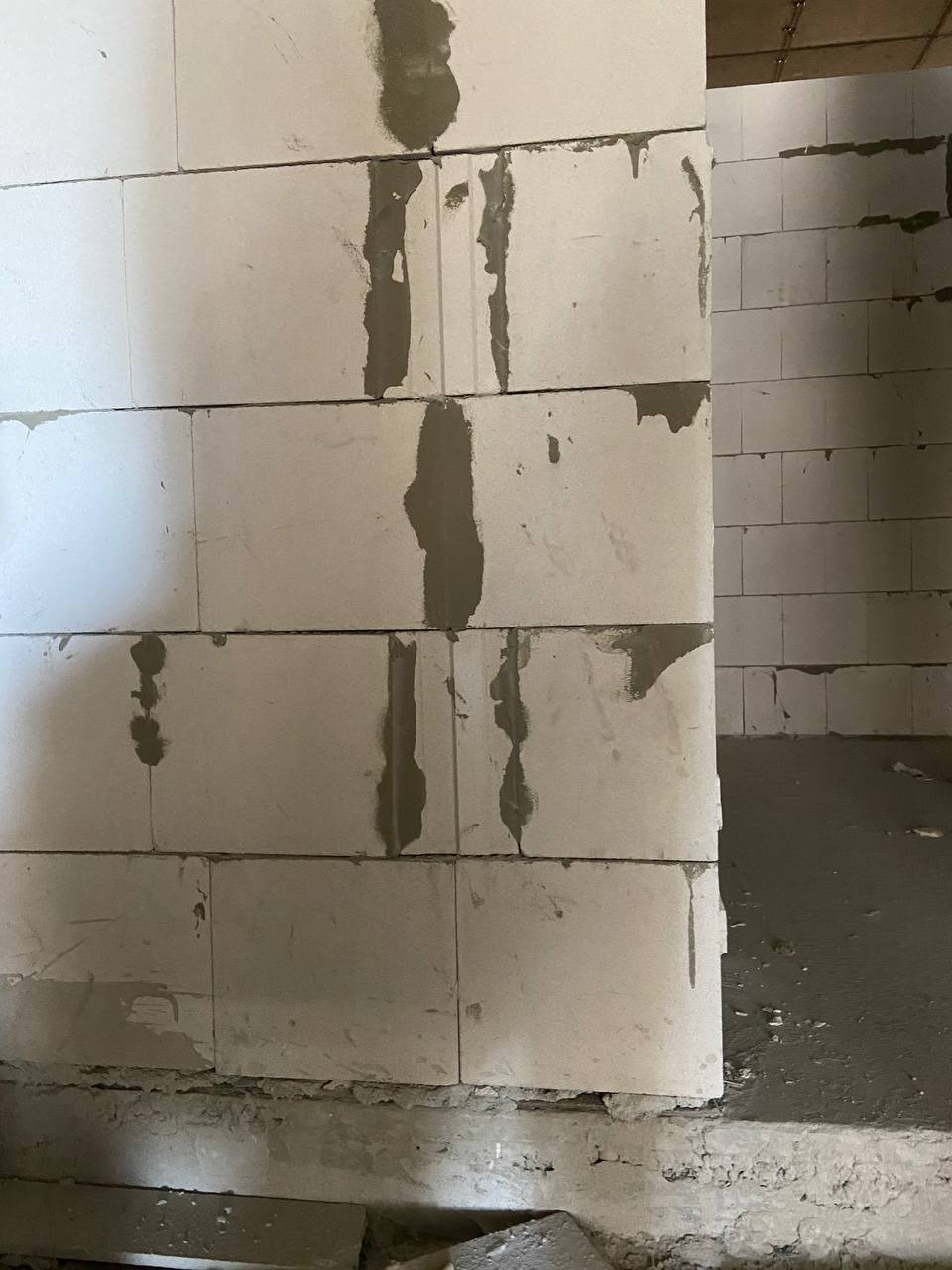 Ściana z białych bloczków, świeżo połączonych zaprawą, z widocznymi pionowymi śladami zaprawy na spoinach, surowy stan wykończenia, widok na wnętrze pomieszczenia w tle.