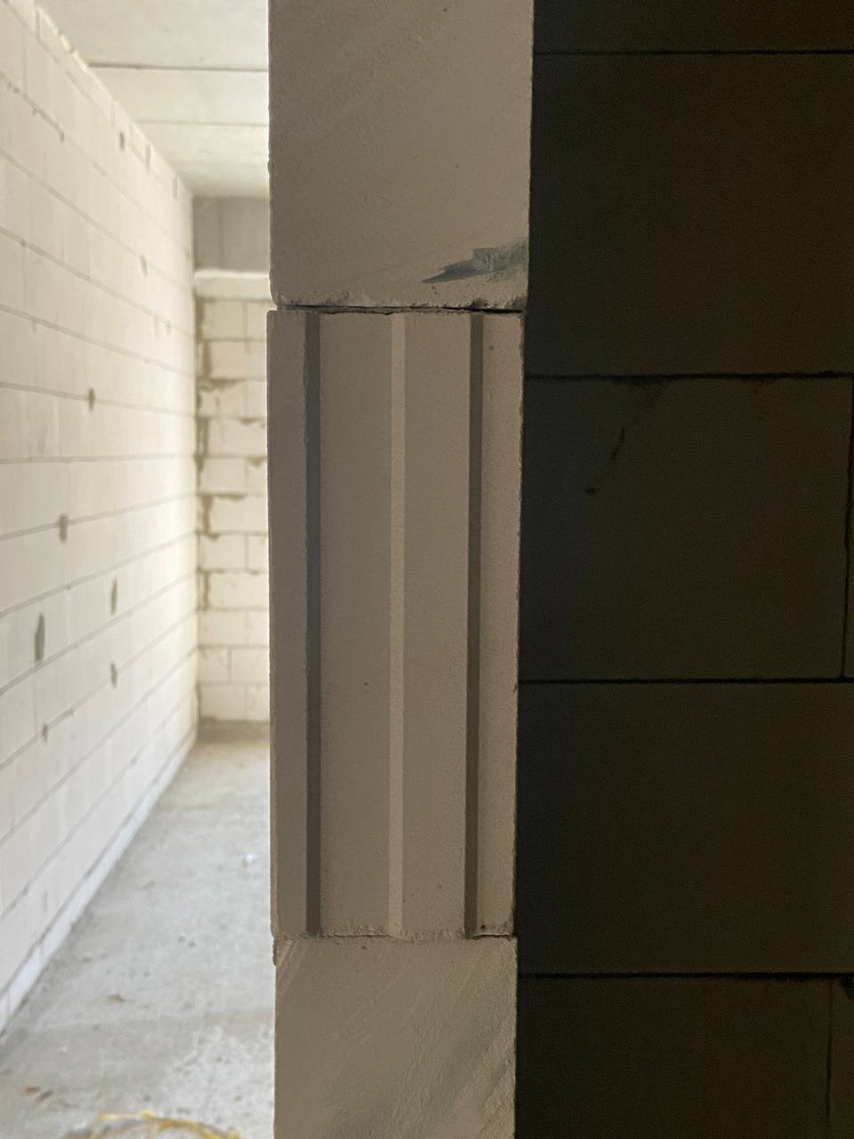Wnętrze budynku w trakcie budowy, widok na korytarz przez otwór w ścianie z szarych i białych bloczków betonowych. Bloczki po lewej stronie mają dekoracyjne pionowe żłobienia.
