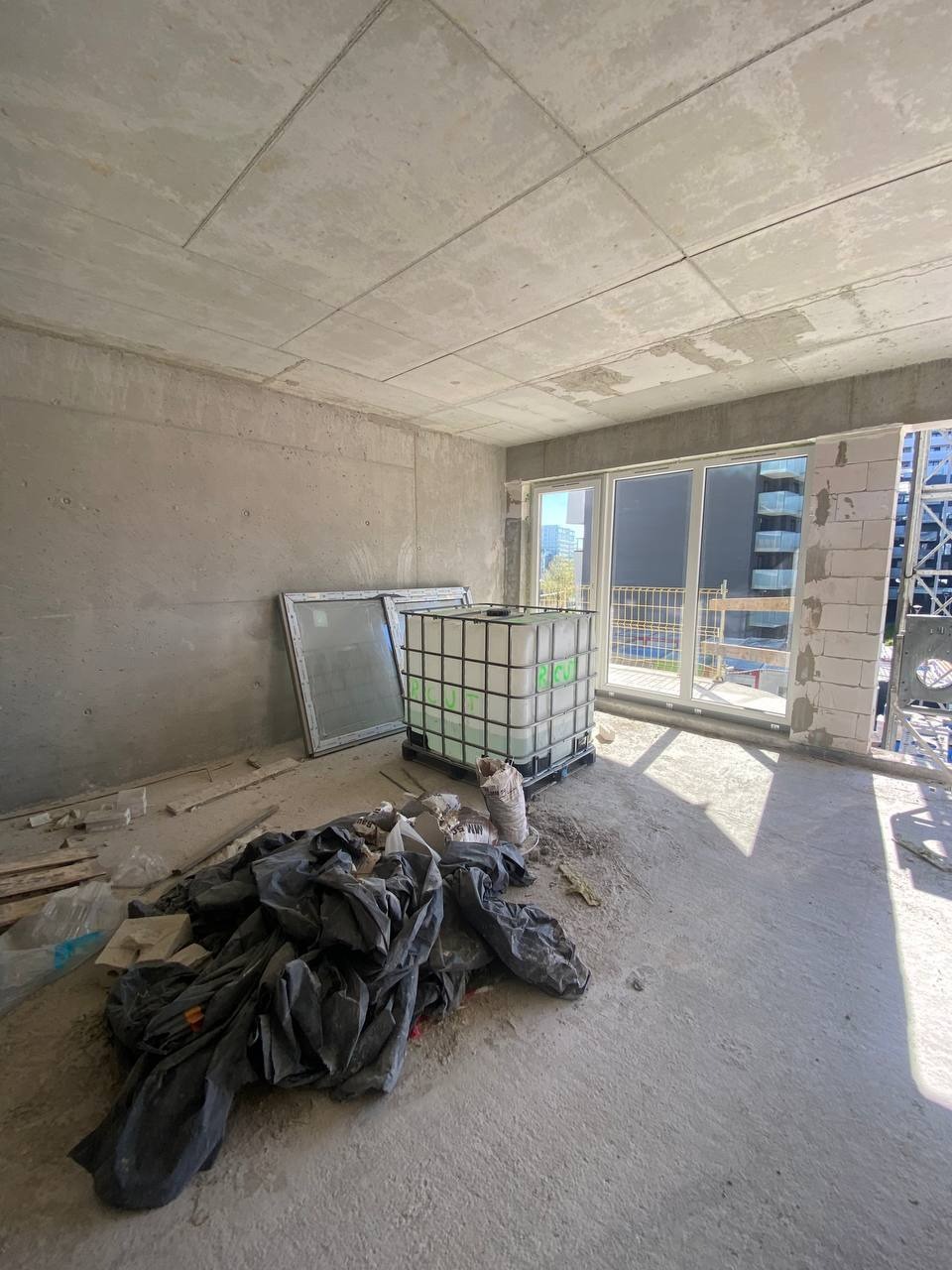 Surowe wnętrze w trakcie budowy z betonowymi ścianami, widoczną instalacją sufitową, oknami balkonowymi, paletą z tworzywa, workami z zaprawą i stertą czarnej folii budowlanej.