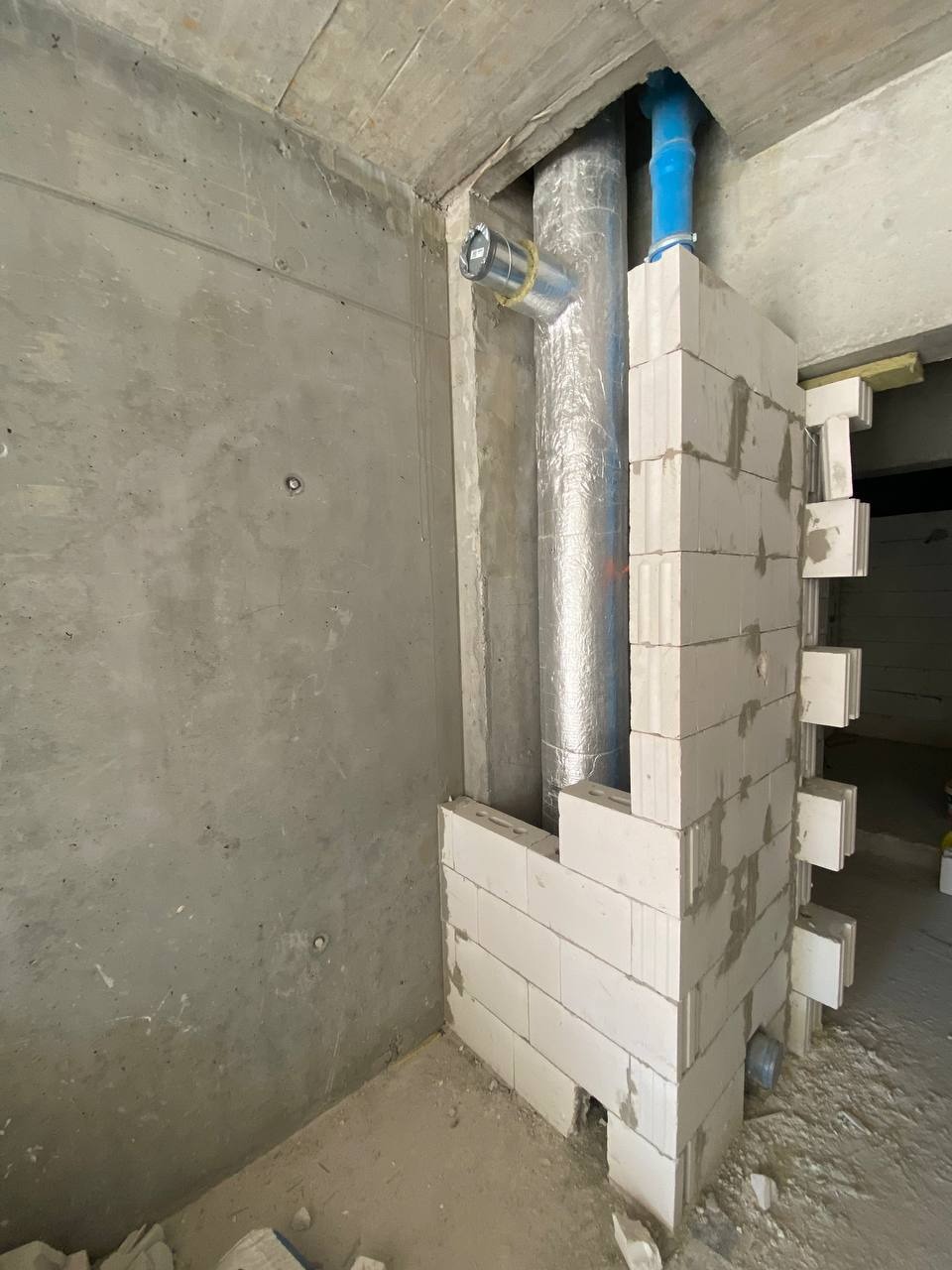 Wznoszenie ścianki działowej z białych bloczków wokół rury wentylacyjnej, zabezpieczonej folią aluminiową, na tle surowej, betonowej ściany w trakcie remontu.