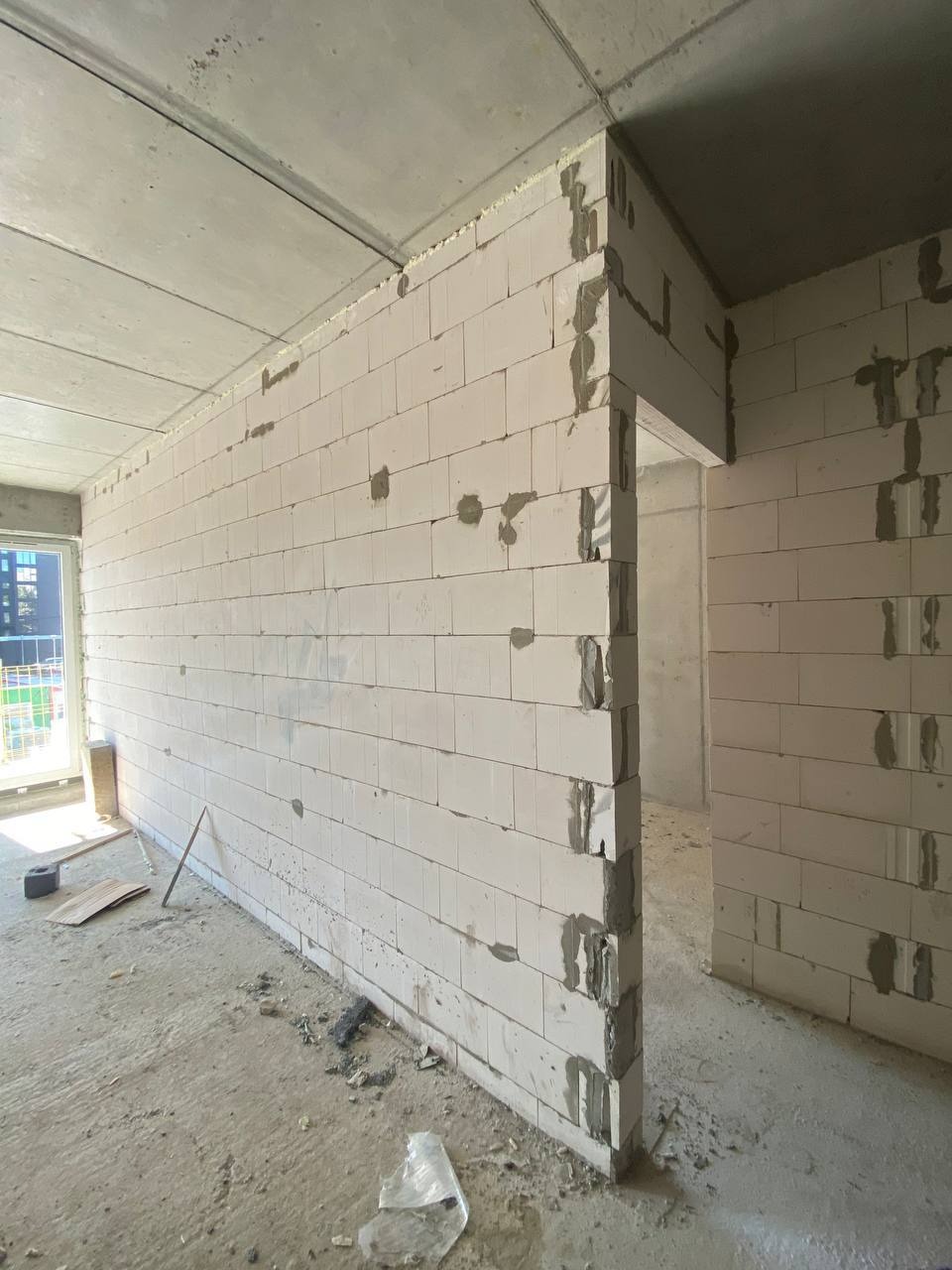 Wnętrze w trakcie budowy z widoczną ścianą z białych bloczków, surowy betonowy strop, fragment okna balkonowego i prowizoryczne narzędzia na podłodze.