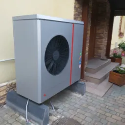 Szara pompa ciepła powietrze-woda na betonowych podstawach przed budynkiem z wejściem wykończonym kamieniem elewacyjnym, widoczne naklejki informacyjne na obudowie.