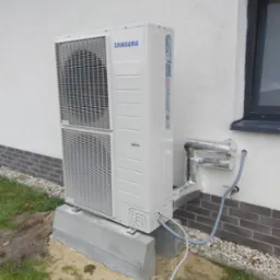 Zewnętrzna jednostka klimatyzacji Samsung zamontowana na betonowym fundamencie przy ścianie budynku, z widocznymi przewodami w izolacji termicznej i elementami instalacji elektrycznej w ziemi.