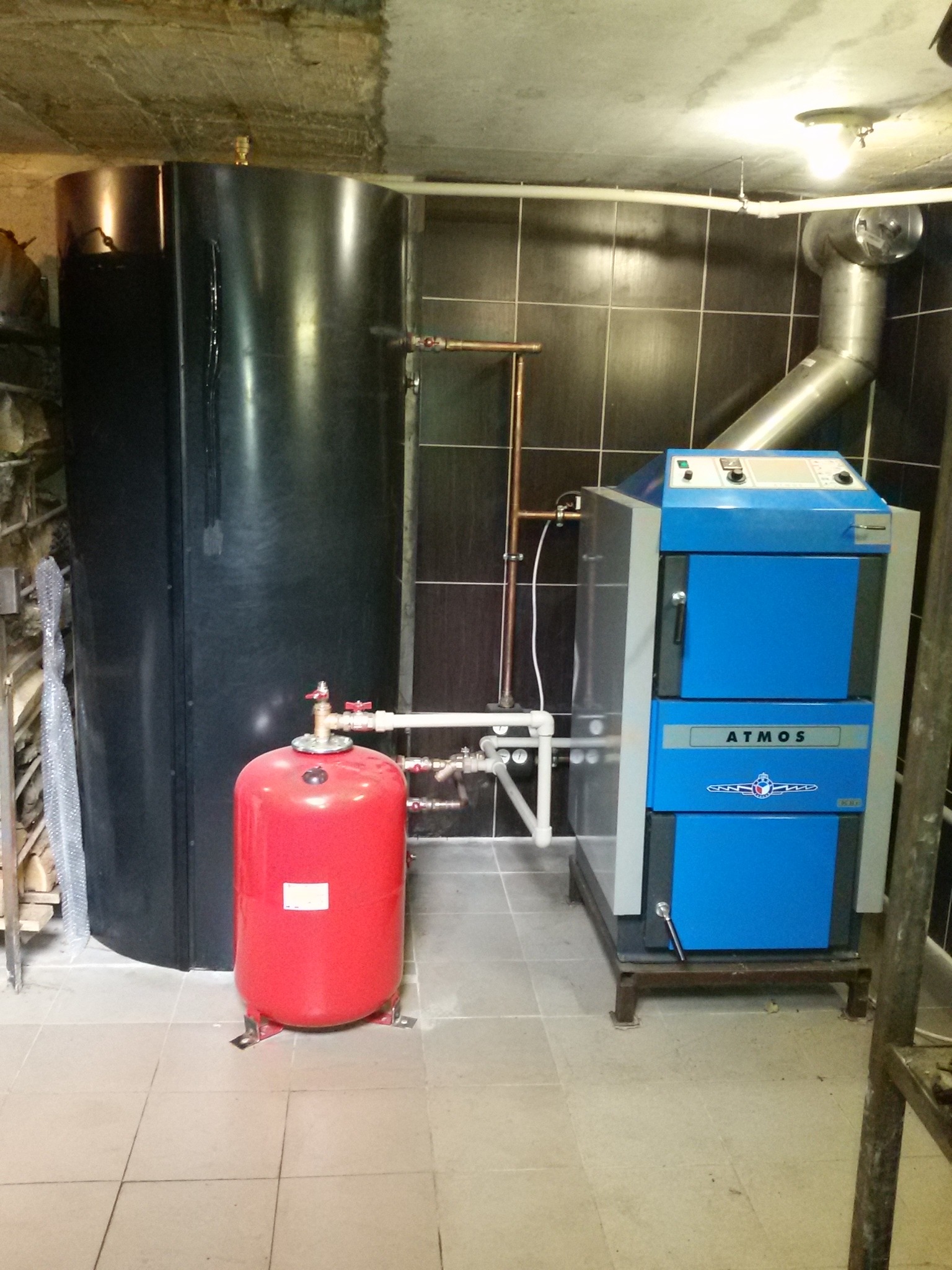 Nowoczesny piec na paliwo stałe Atmos w kotłowni, z czarnym zbiornikiem akumulacyjnym i czerwonym naczyniem przeponowym, rury miedziane, płytki ceramiczne na ścianach.