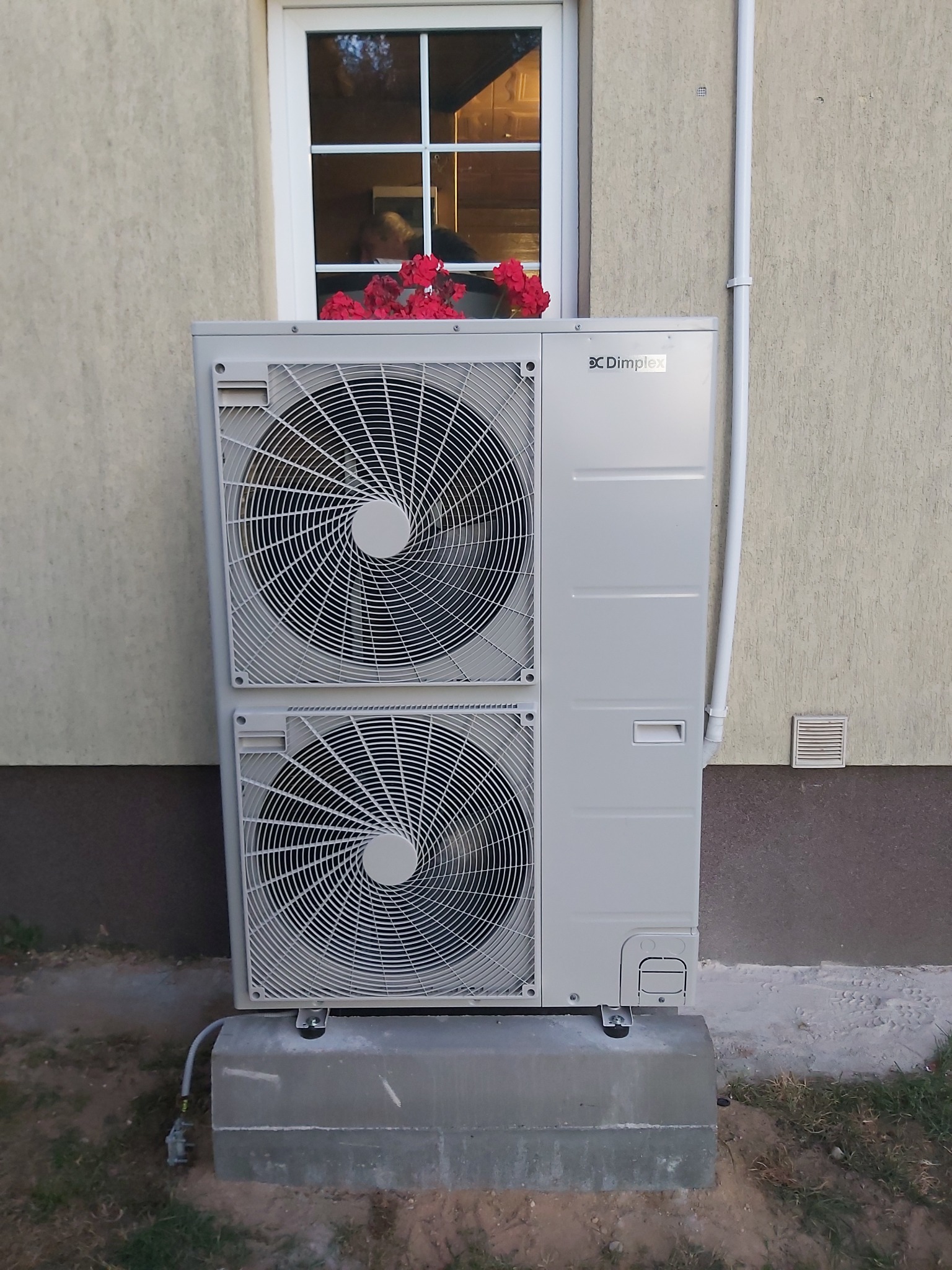 Zewnętrzna jednostka pompy ciepła Dimplex zamontowana na betonowym fundamencie przy ścianie budynku z oknem, nad jednostką czerwone kwiaty w doniczce.