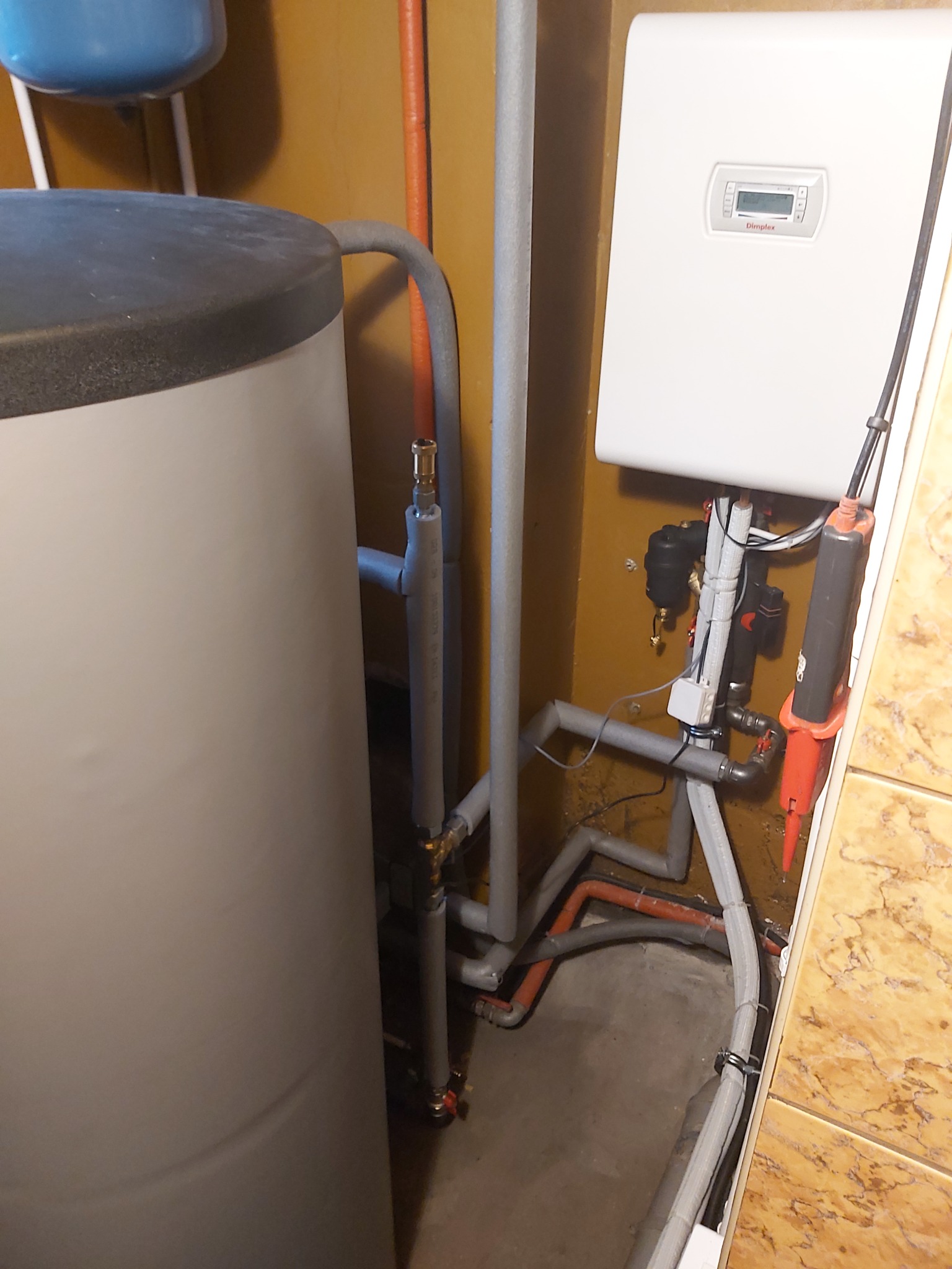 Bojler z podłączonymi rurami, zaworami i białym panelem sterowania Dimplex na żółtej ścianie wyłożonej płytkami.