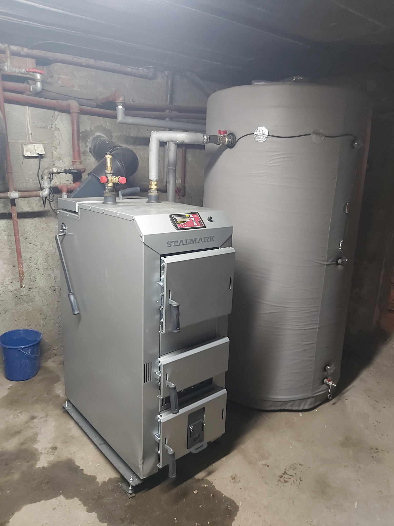 Szary piec z podajnikiem na paliwo stałe marki Stalmark, z podłączonym zasobnikiem wody, zainstalowany w piwnicy z betonową posadzką i widocznymi rurami instalacji grzewczej.