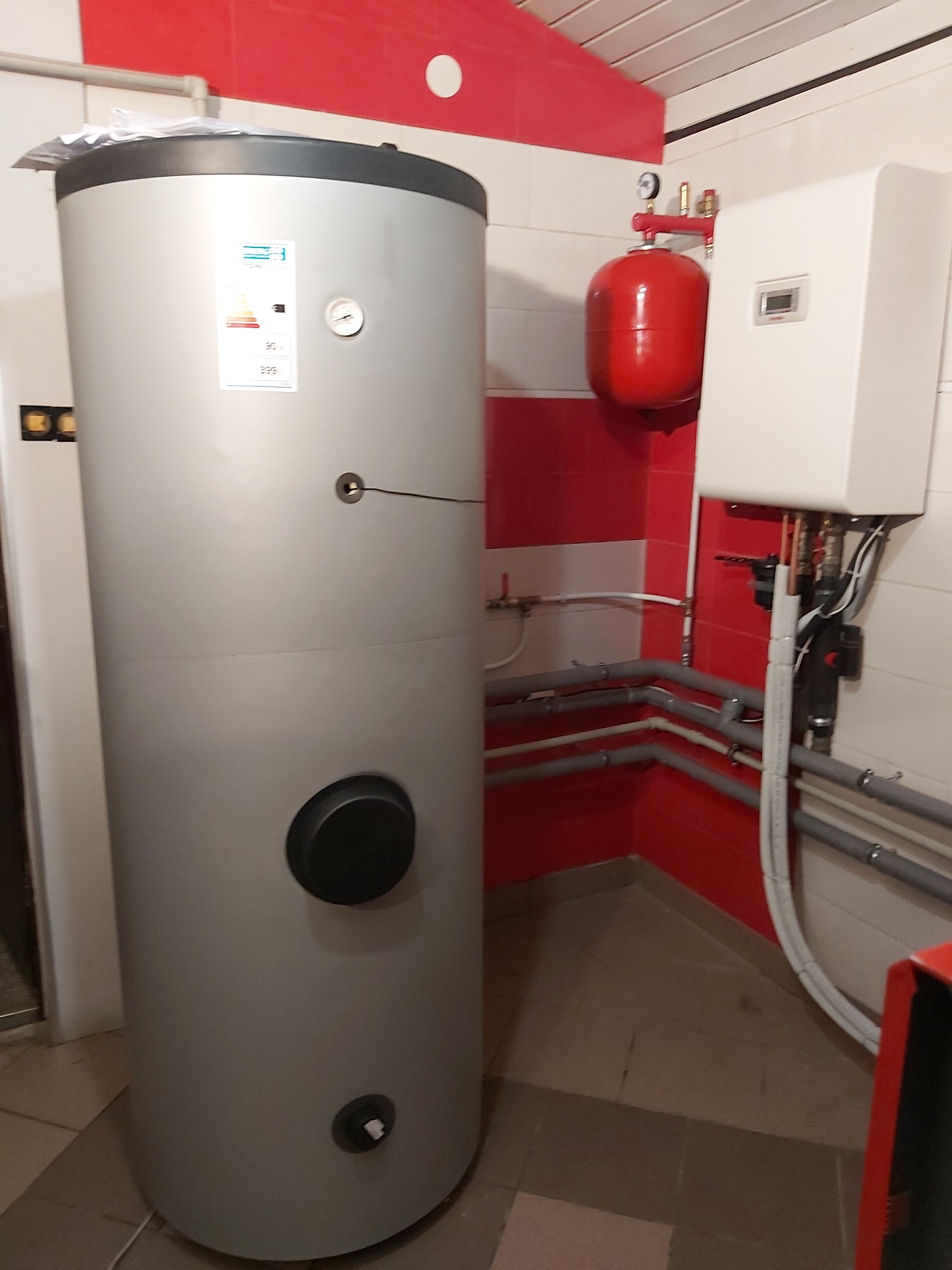 Szary, cylindryczny zbiornik na wodę z panelem sterowania obok, podłączony do instalacji hydraulicznej w pomieszczeniu z czerwono-białymi ścianami.