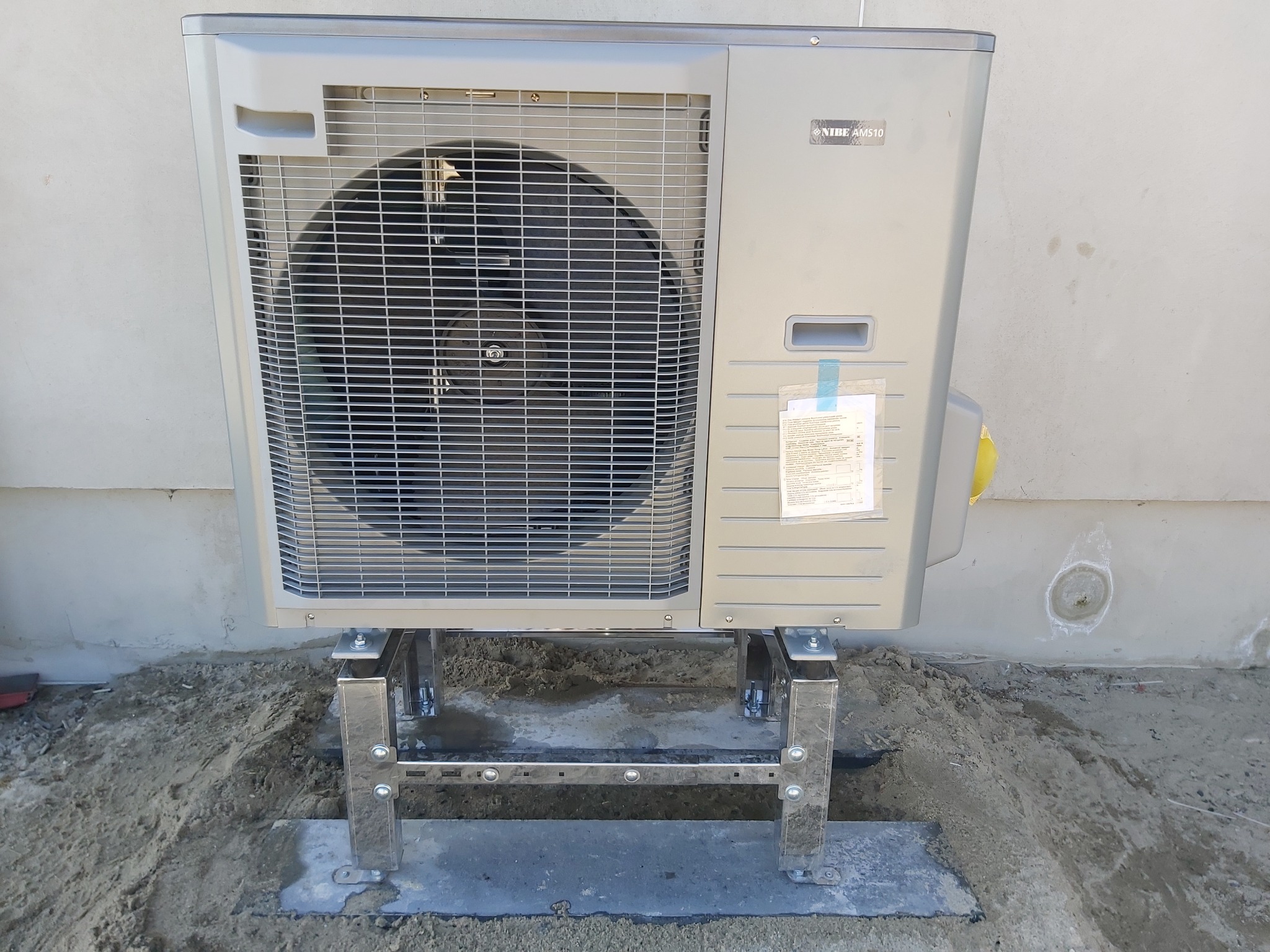 Zewnętrzna jednostka pompy ciepła Nibe AMS10 zamontowana na metalowej konstrukcji wsporczej, z widocznym podłożem z piasku i betonu oraz fragmentem ściany budynku.