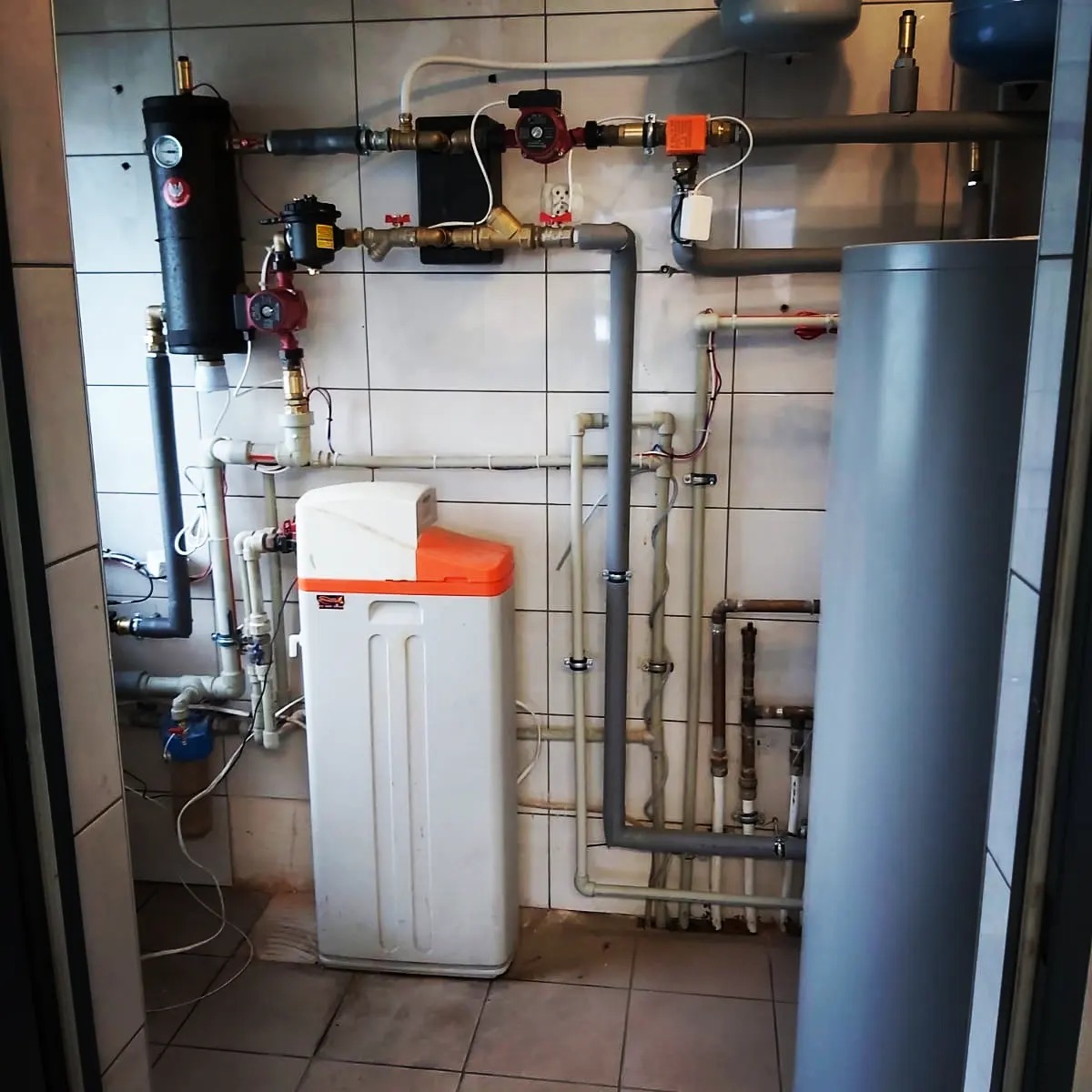 Instalacja hydrauliczna w kotłowni z widocznym zmiękczaczem wody z pomarańczową pokrywą, zbiornikiem wyrównawczym i licznymi rurami na tle białych płytek.