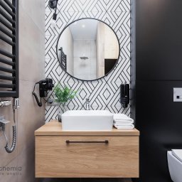 Studio Archemia - Nowoczesna łazienka z drewnianą szafką pod umywalką, okrągłym lustrem i geometryczną mozaiką na ścianie, uzupełniona czarnym grzejnikiem drabinkowym i zabudową.