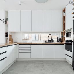 Studio Archemia - Nowoczesna kuchnia z białymi szafkami i drewnianym blatem, czarną lodówką i designerską lampą wiszącą, widok na okno z czarną armaturą.