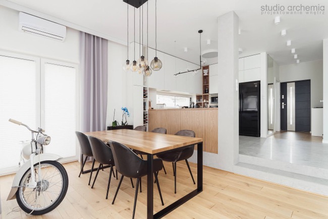 Jasna jadalnia w nowoczesnym stylu z drewnianym stołem na metalowych nogach, czarnymi krzesłami, wiszącymi lampami z żarówkami Edisona i białym motocyklem przy oknie, w tle widoczna kuchnia...