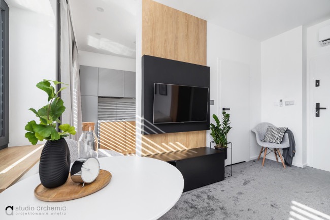 Nowoczesny salon z okrągłym białym stołem, czarnym wazonem z zielonymi liśćmi, telewizorem na drewnianej ścianie i szarym dywanem.