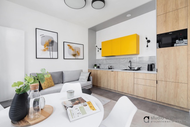 Nowoczesne wnętrze mieszkania z żółtymi szafkami kuchennymi, szarą sofą, okrągłym białym stołem i dekoracyjnymi obrazami na ścianie.