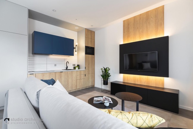 Nowoczesne wnętrze salonu z aneksem kuchennym w Krakowie. Widoczna sofa, stoliki kawowe, szafki kuchenne w odcieniach drewna i granatu, ściana TV z podświetleniem LED.