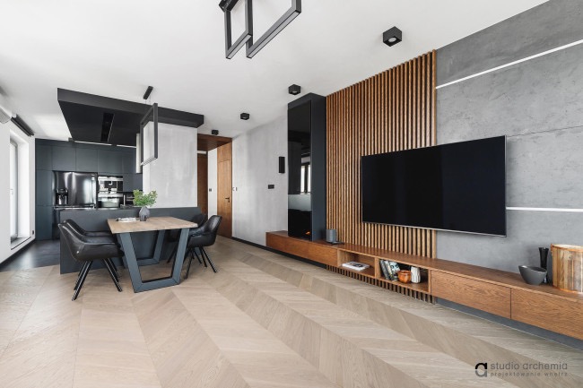 Nowoczesne wnętrze mieszkania z drewnianą ścianą telewizyjną, stołem jadalnym z czarnymi krzesłami i aneksem kuchennym w ciemnych kolorach.